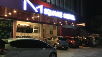 Ein Hotel Eingang mit einem auffälligen 'M'-Schild, ein Parkplatz mit mehreren Autos und eine sichtbare Treppe zum Eingang.