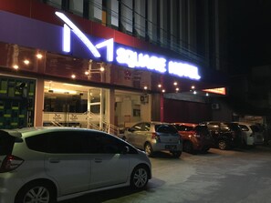 A hotel entrance with a prominent 'M' sign, a parking area with several cars, and a visible staircase leading to the entrance. - Msquare Hotel (Palembang)