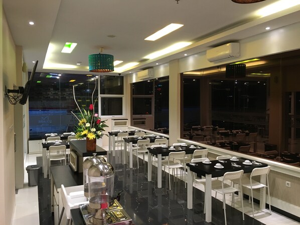 A restaurant with a long counter, white chairs, and a coffee machine. - Msquare Hotel (Palembang)