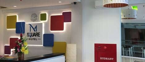 Zona de recepción de un hotel con diseño moderno, que incluye un mostrador de recepción, un reloj y un cartel que dice "SQUARE HOTEL". Hay espacios de descanso con bancos y otomanas, además de un armario de hidrantes rojo.