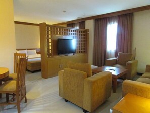 Quarto de hotel com televisão sobre suporte de madeira, um sofá, uma cadeira e uma pequena mesa.
