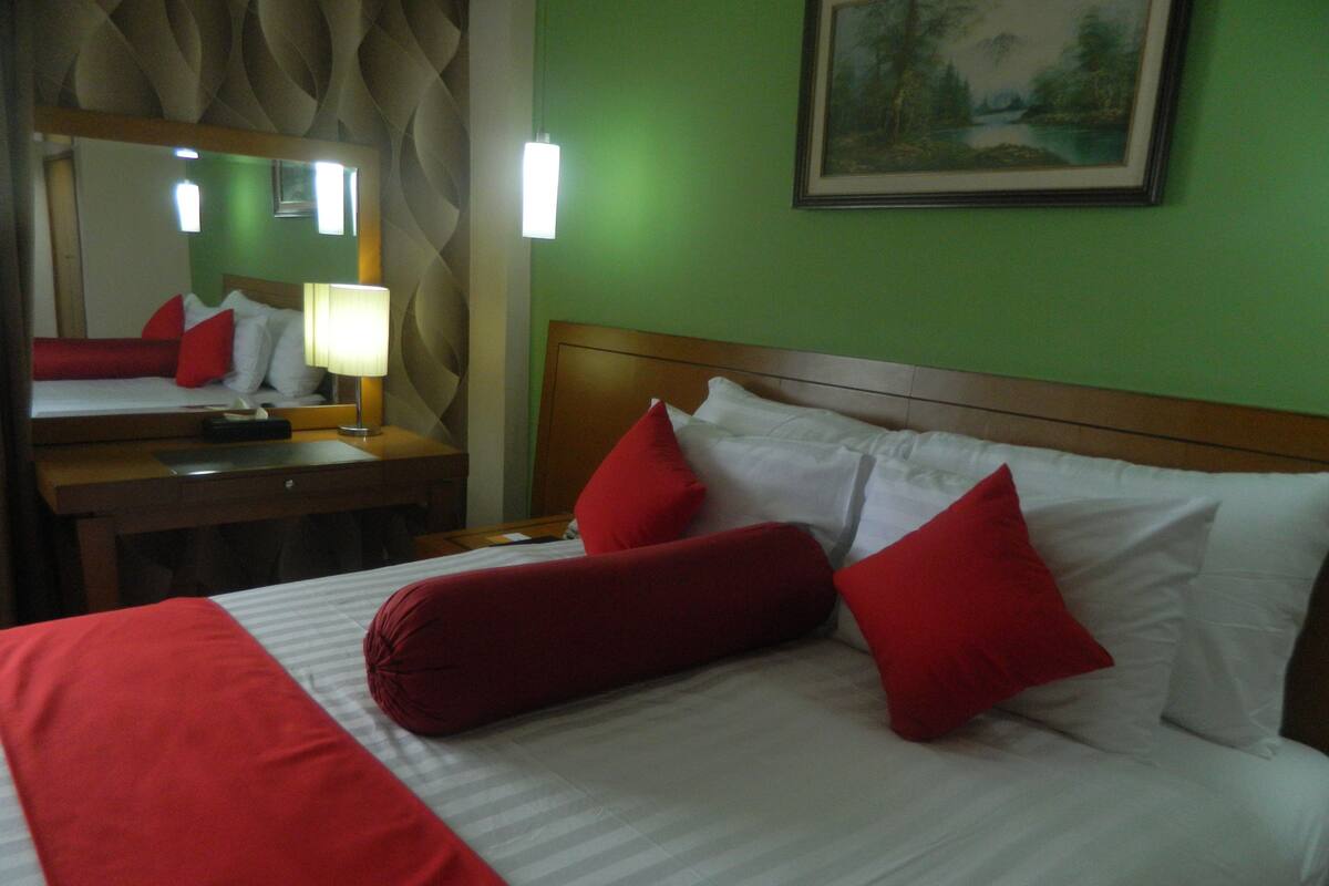 Habitación de hotel con cama, mesita de noche, lámpara, espejo y un cuadro en la pared.