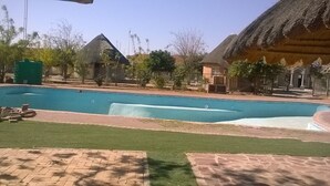 A swimming pool with a grassy area in the foreground, surrounded by buildings with thatched roofs. - Sahara Stones Hotel (Palapye)