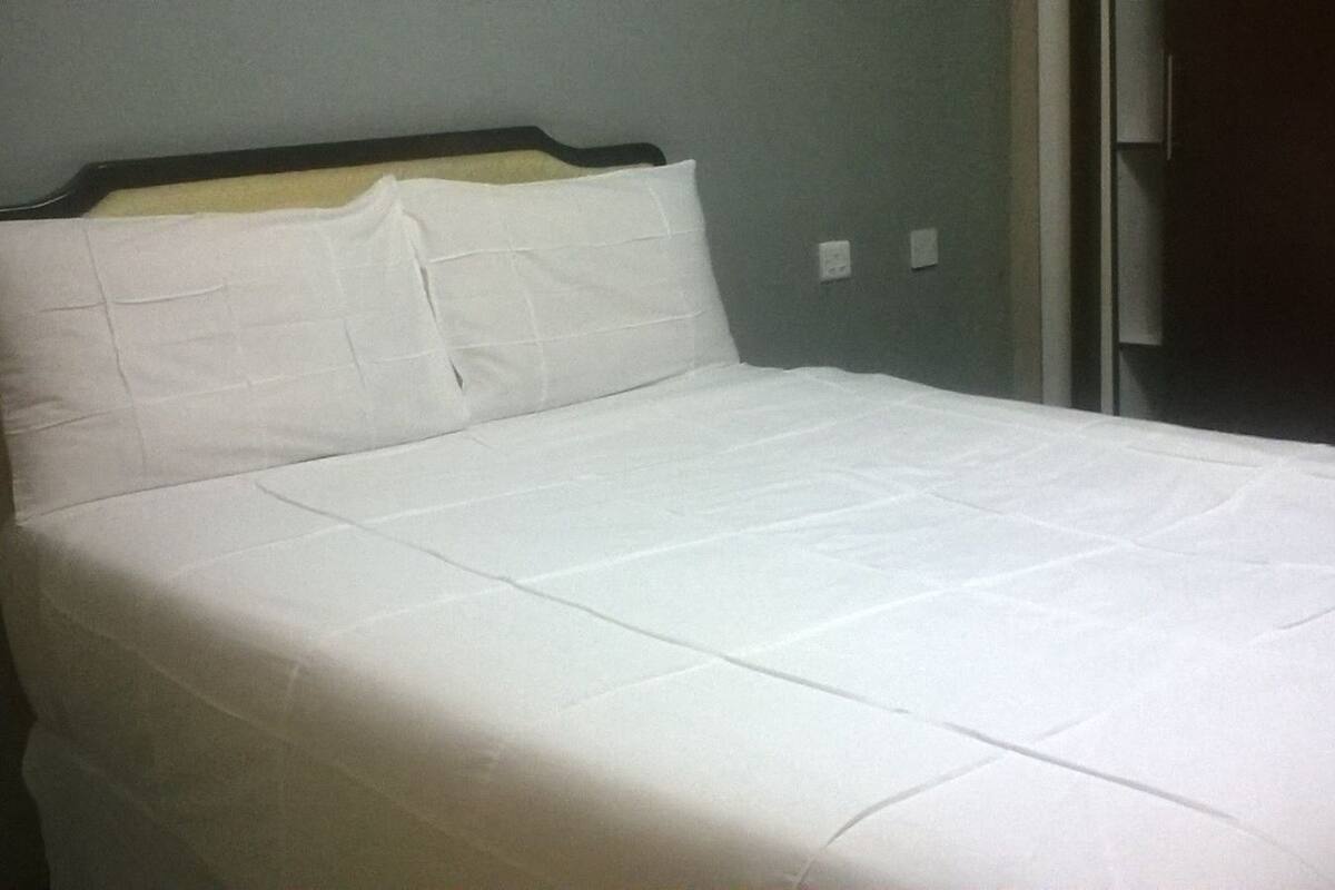 A plain white bed with a wooden headboard in a room with light-colored walls and a door to the right.