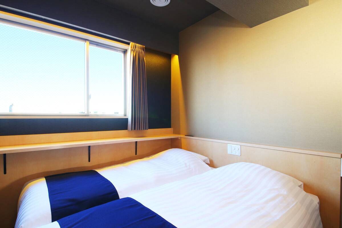 Een hotelkamer met een bed, een raam met gordijnen, een plank en een stopcontact.