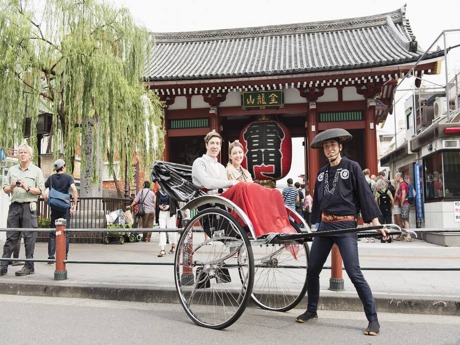 A traditional building with a tiled roof and wooden beams, a person in traditional attire pushing a covered carriage, and pedestrians in the background.