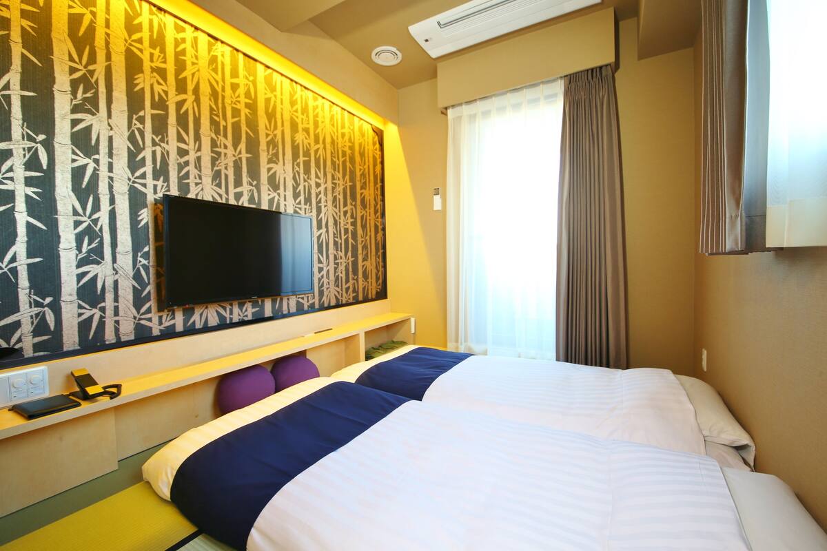 Een hotelkamer met twee bedden, een flatscreen-tv bevestigd aan een wand met bamboemotief, een raam met gordijnen en een plafondairconditioner.