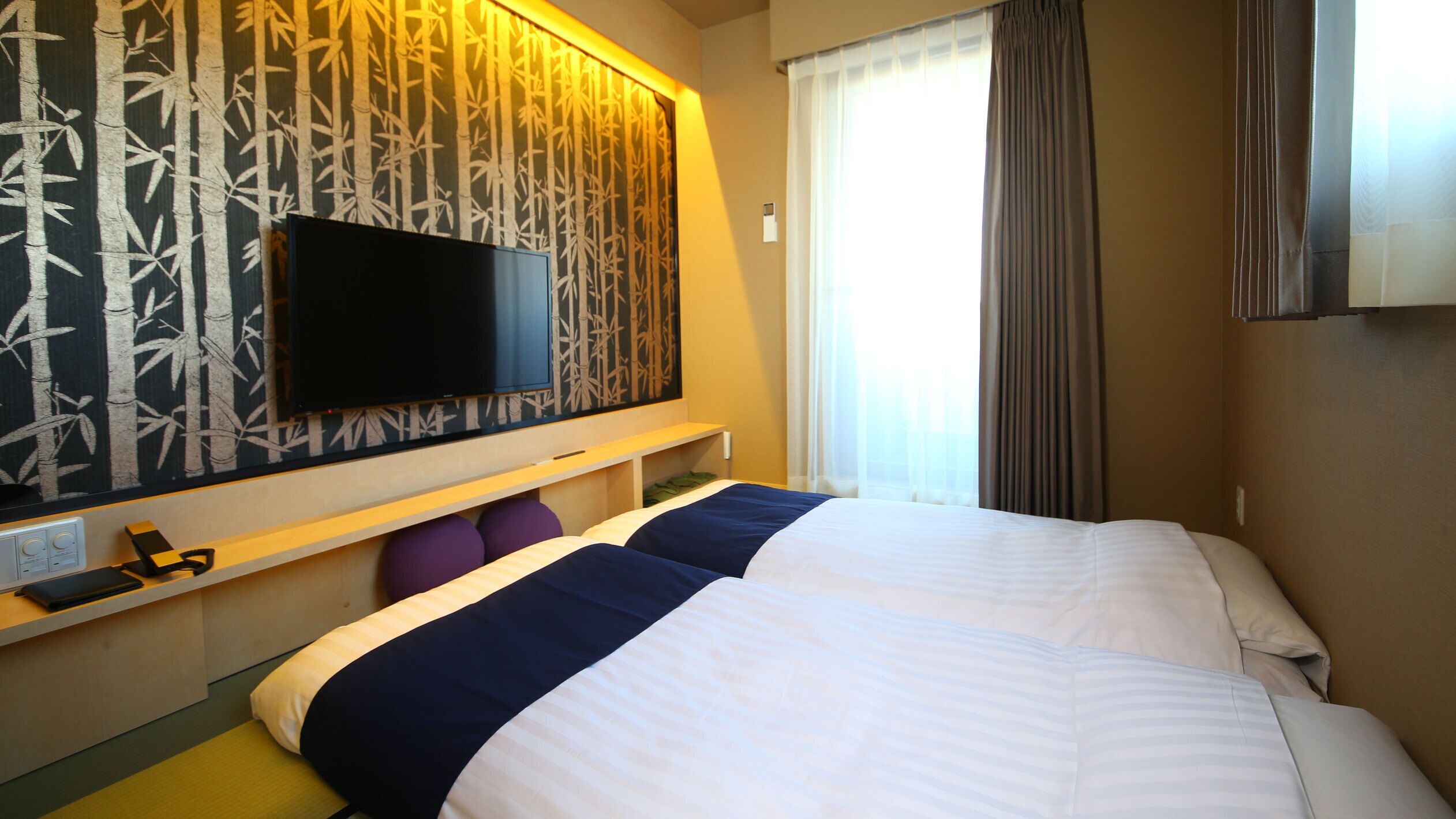 Une chambre d’hôtel avec un lit, un téléviseur fixé au mur, une fenêtre avec des rideaux et un mur orné d’un motif en bambou.