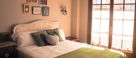 Una cama bien tendida con una manta verde y almohadillas decorativas, una mesita de noche con una lámpara y una pared con cuadros enmarcados.