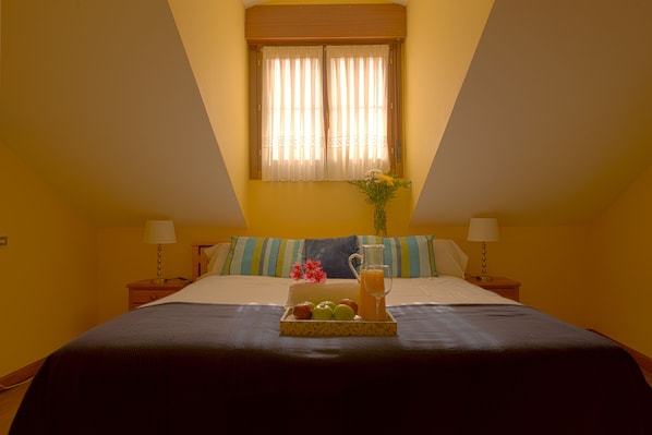 Hotelkamer met een bed, nachtkastje met een lamp, raam met gordijnen en dienblad met fruit en een drankje.