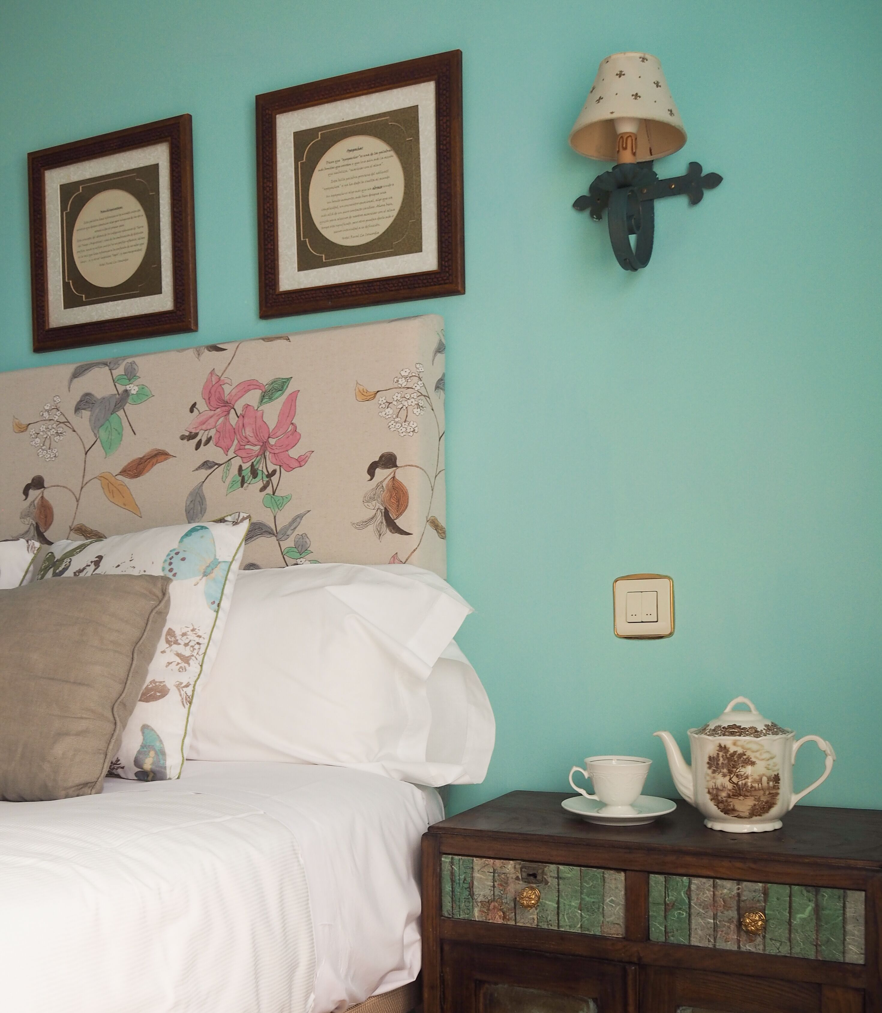 A bedroom with a bed, a nightstand with a teapot and cup, and two framed certificates on the wall.