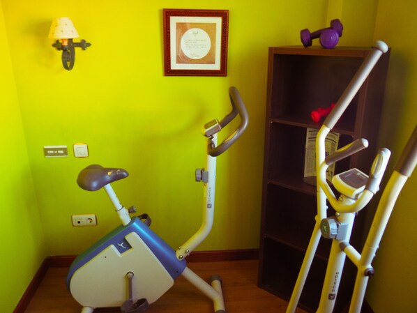 A room with a stationary exercise bike, a wall-mounted lamp, and a shelf with dumbbells.