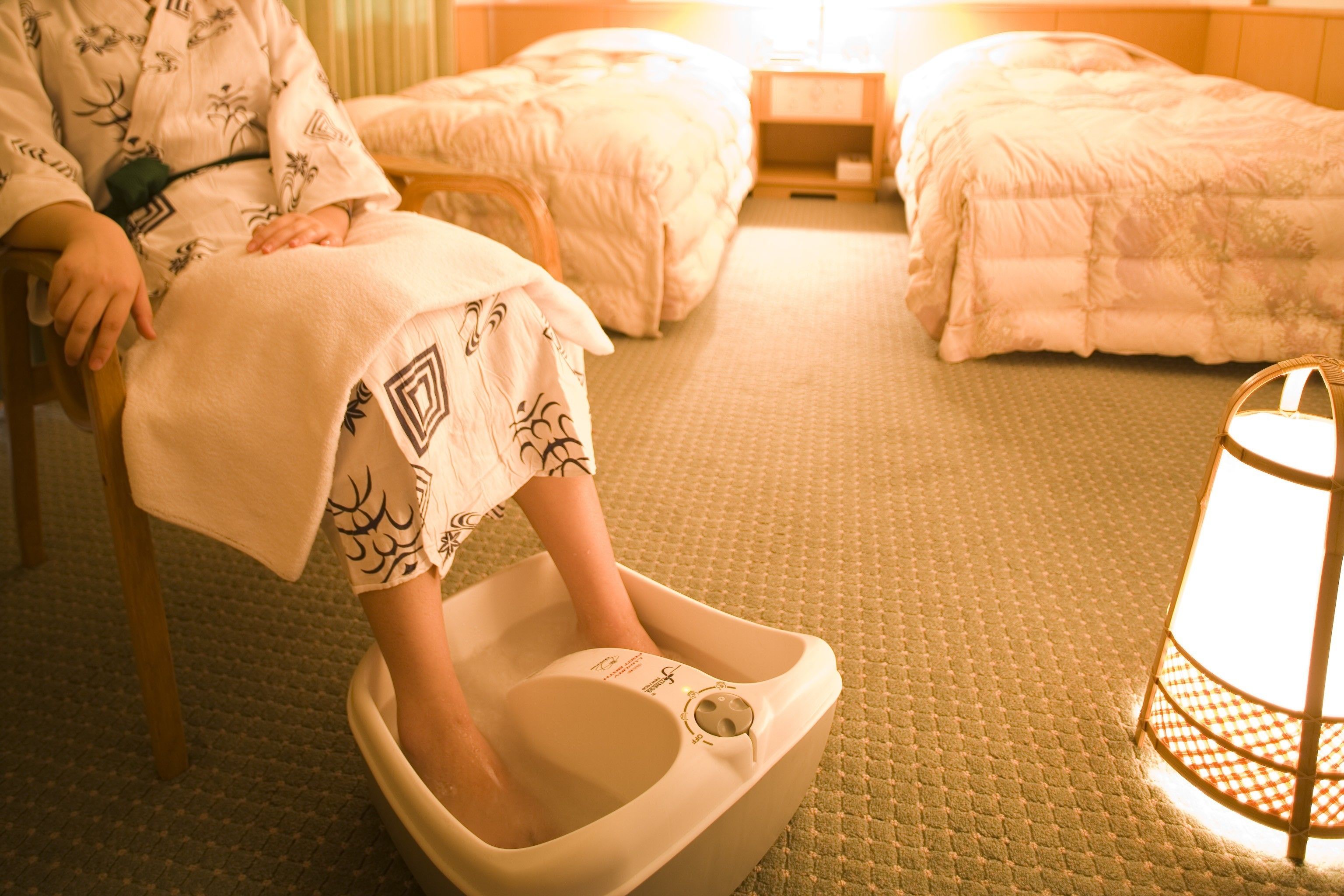 Une personne est assise dans un bain de pieds, dans une pièce où se trouvent deux lits et une table de chevet.