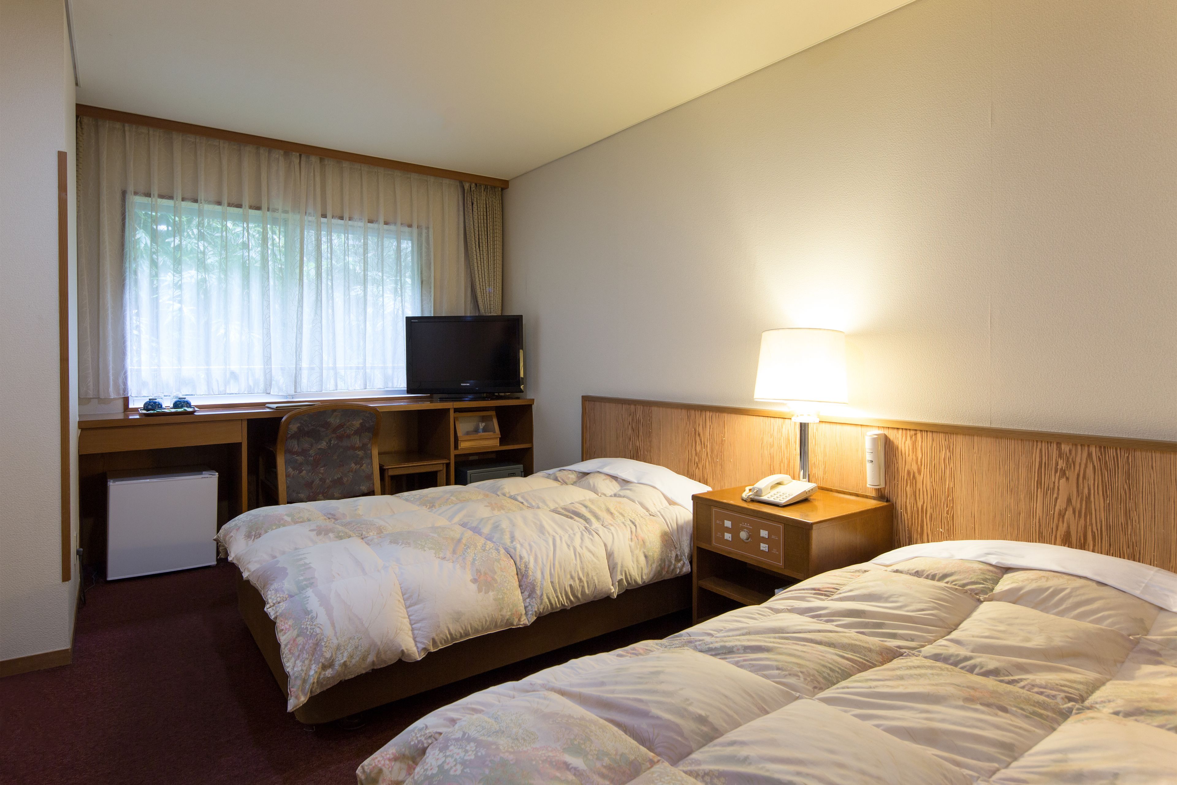 A hotel room with two beds, a desk with a television, a lamp, and a refrigerator.