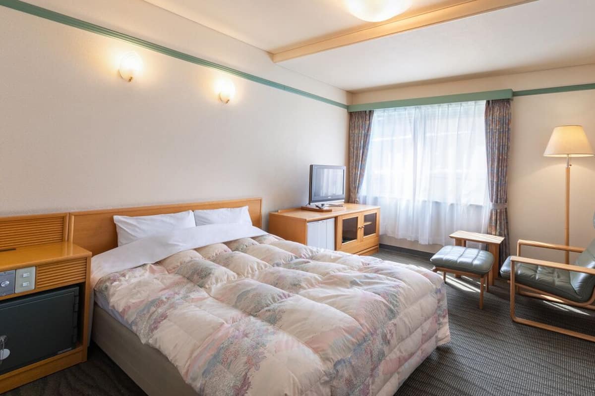 A hotel room with a bed, a desk with a television, a chair, a lamp, and a window with curtains.