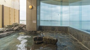 Un onsen japonés tradicional con una bañera de piedra rodeada por un muro de piedra, una valla de madera y un recinto de vidrio con vistas al mar.