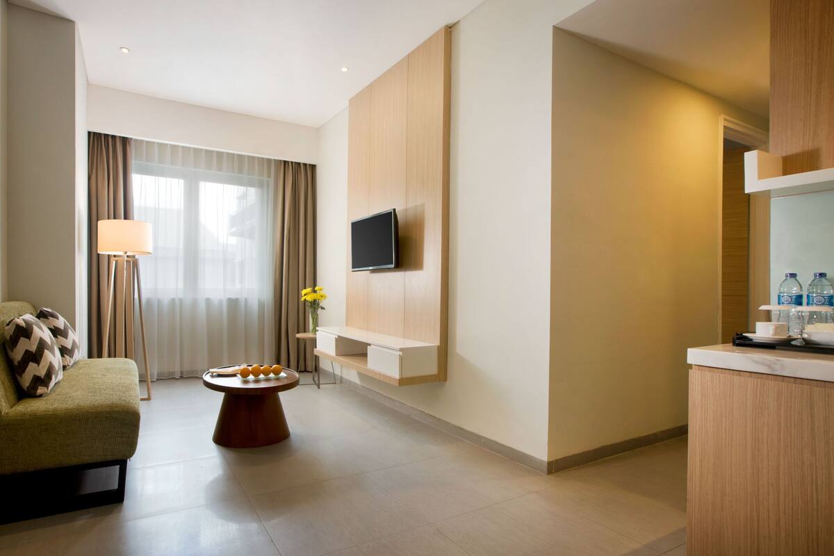 A modern hotel room with a sofa, a small round table, a flat-screen TV mounted on the wall, and a kitchenette area.