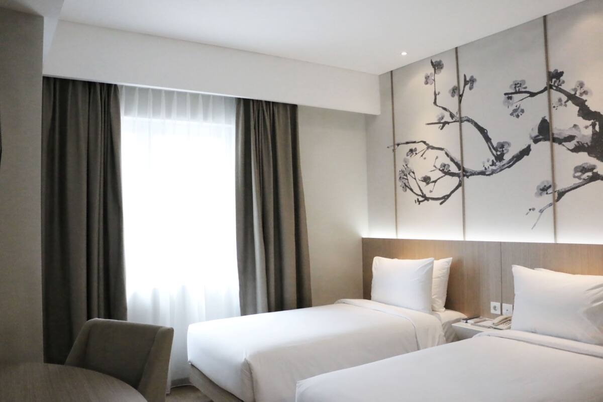 Une chambre d’hôtel avec deux lits, une grande fenêtre avec des rideaux, une œuvre d’art accrochée au mur et une chaise.
