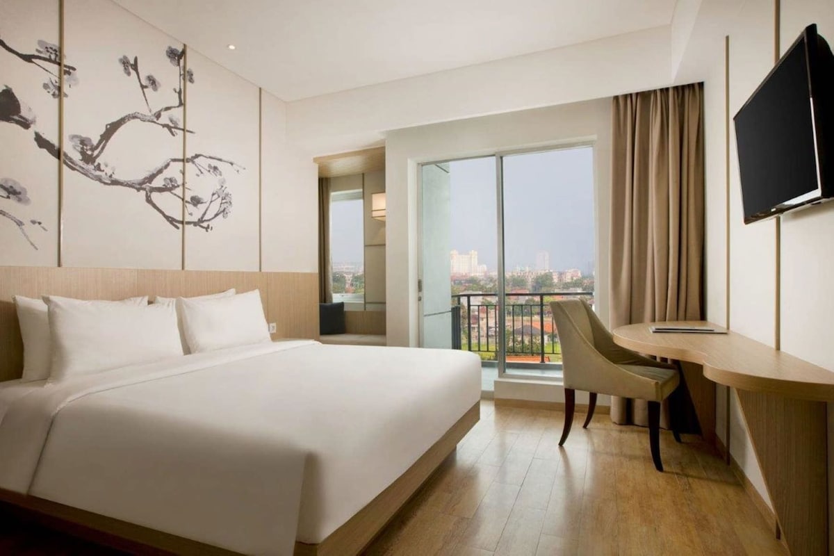 Ein Hotelzimmer mit einem großen Bett, einem Schreibtisch, einem Sessel, einem Fernseher und einem Balkon mit Blick auf die Stadt.