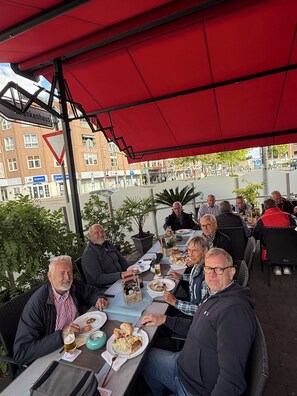 A group of people dining outdoors at a table with a red canopy, surrounded by buildings and greenery.