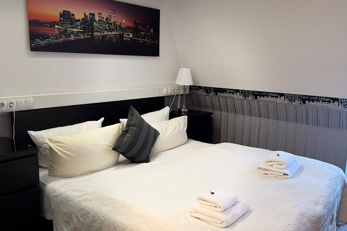 A neatly made bed with pillows, a cityscape painting above, a lamp, and a wall with a city-themed design.