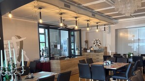 A modern dining area with wooden tables and black chairs, a kitchen in the background, and a chandelier.
