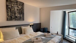 A hotel room with a large bed, a headboard with a geometric pattern, a wall-mounted artwork, a bedside table with a lamp, and a large window with a view of greenery outside.