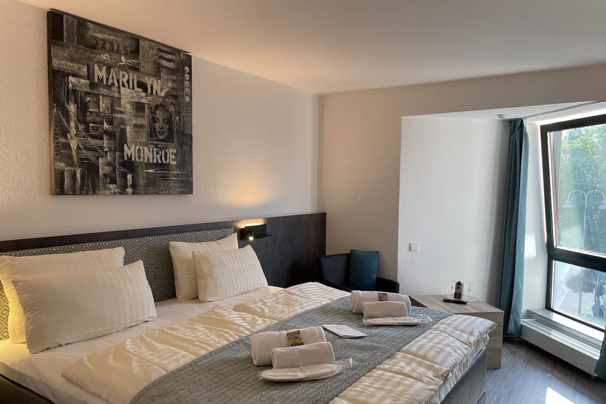 A hotel room with a large bed, a headboard with a geometric pattern, a wall-mounted artwork, a bedside table with a lamp, and a large window with a view of greenery outside.