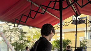 An outdoor seating area with tables and chairs, a person serving a drink, and a building in the background.