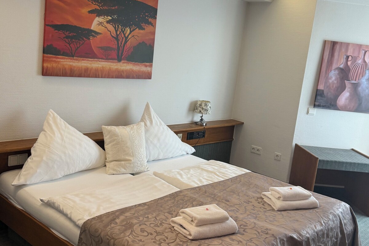 A hotel room with a bed, pillows, a nightstand, a lamp, and two paintings on the walls.