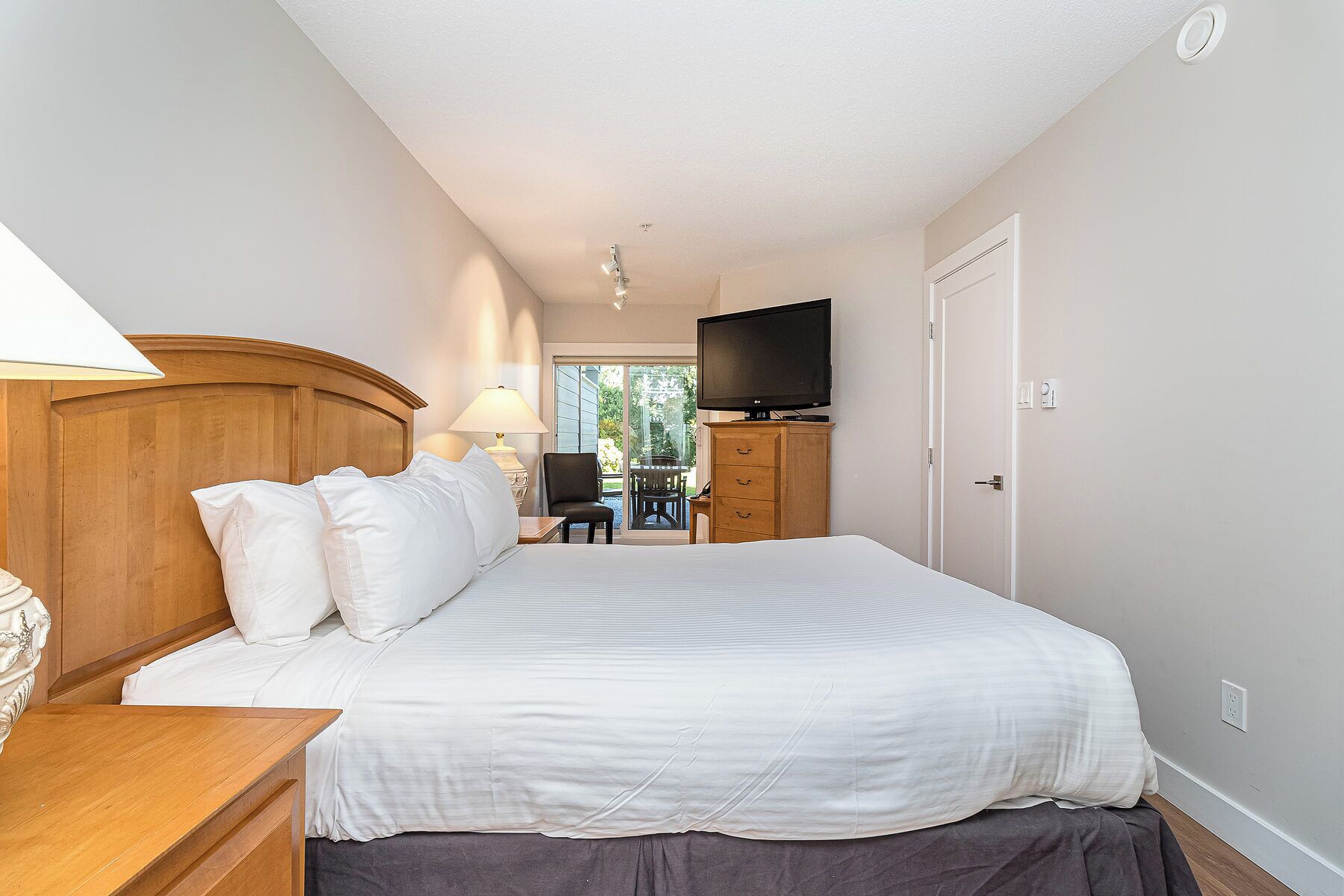 A hotel room with a large bed, a wooden headboard, a flat-screen TV, and a view of the outdoors through a glass door.