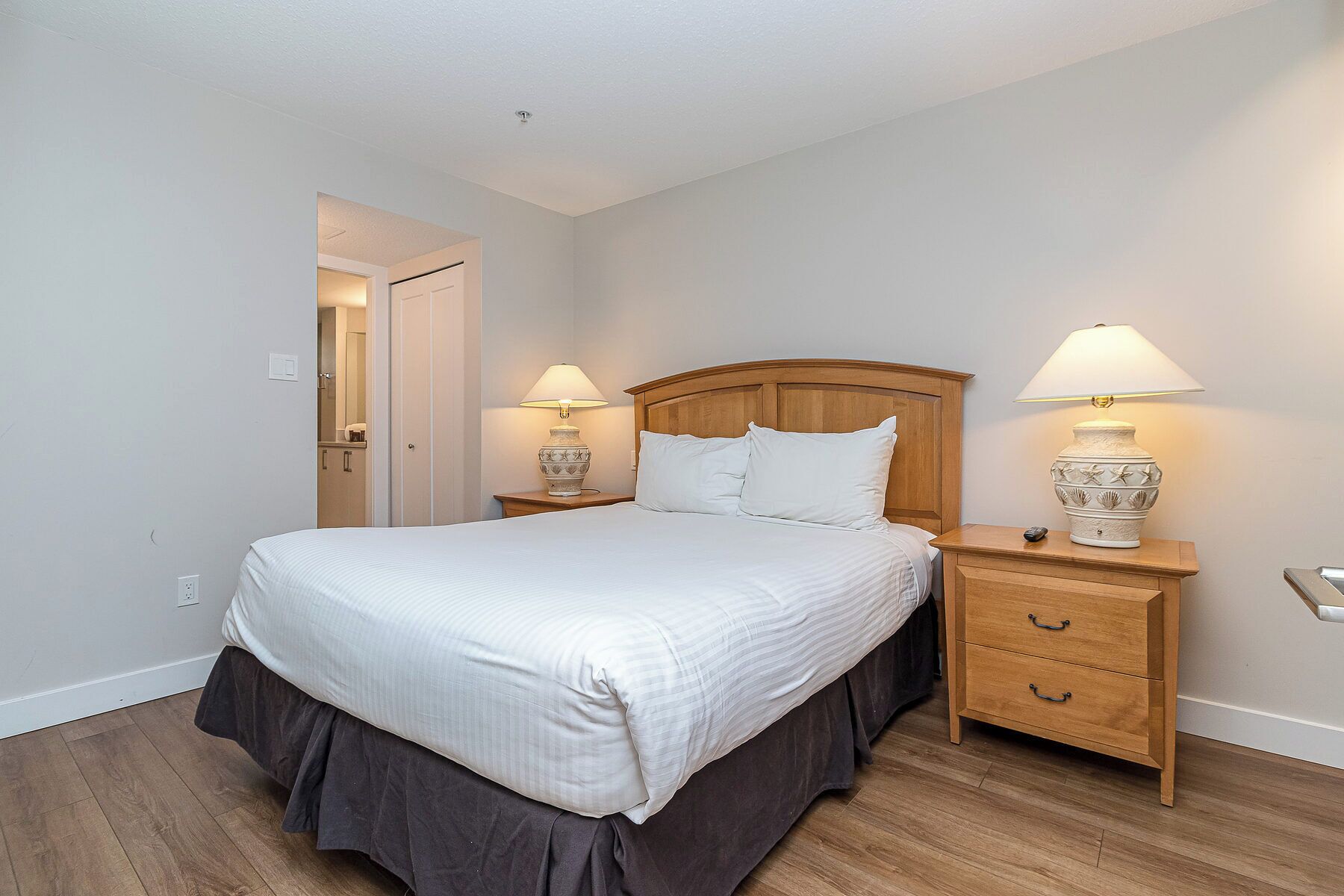 Una cama bien hecha con cabecera de madera, dos mesitas de noche con lámparas y una cómoda de madera con cajones.