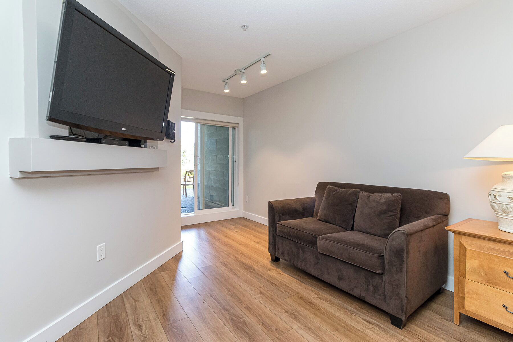 A living room with a flat-screen TV mounted on the wall, a brown sofa, a wooden side table, and a lamp.