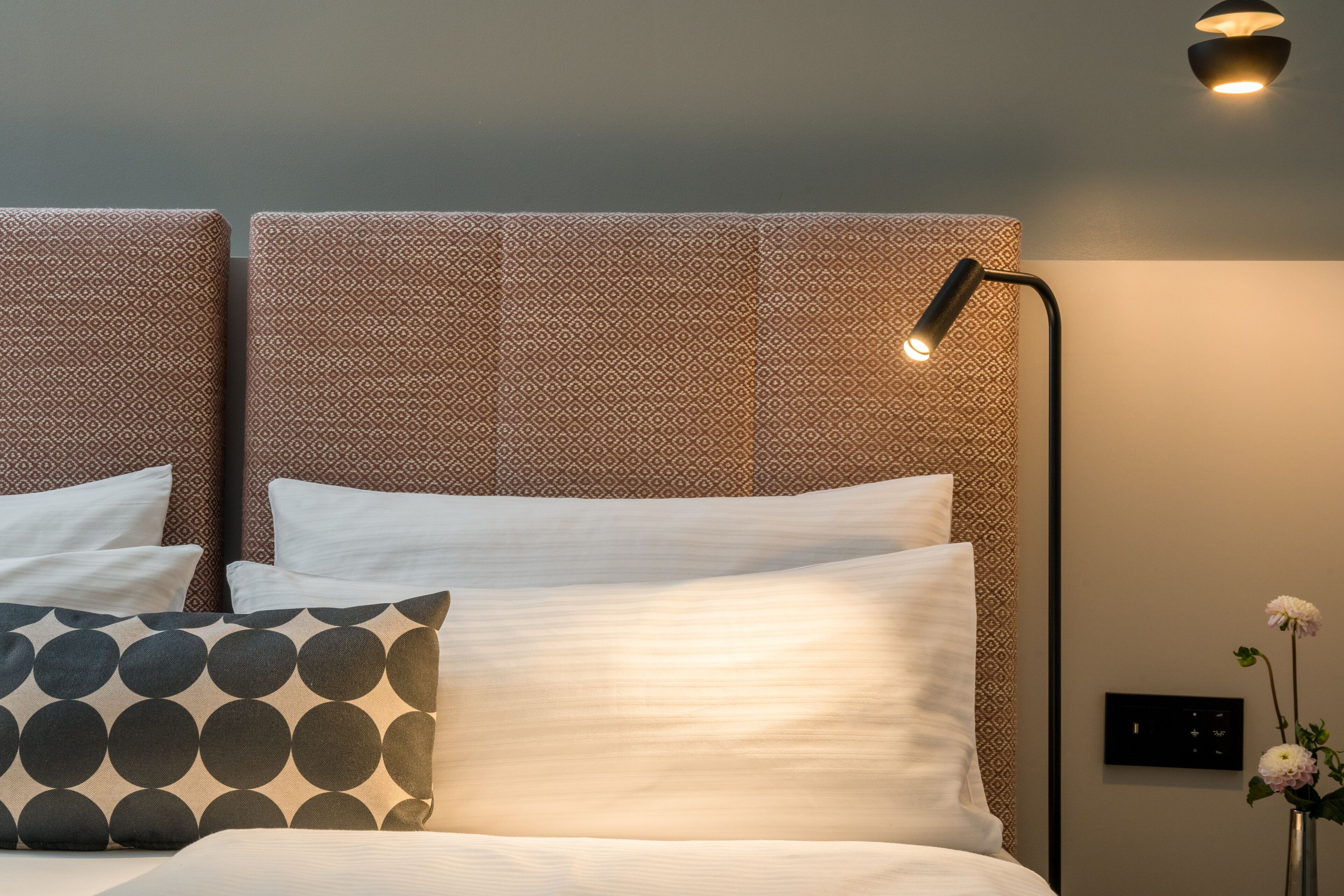A neatly made bed with white and patterned pillows, a headboard with a geometric pattern, and a wall-mounted light fixture.