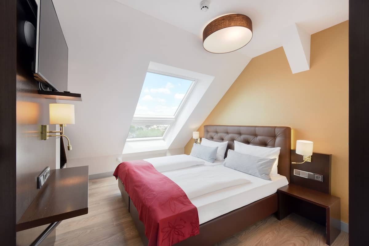 A modern hotel room with a large bed, a desk, a TV, and a skylight.