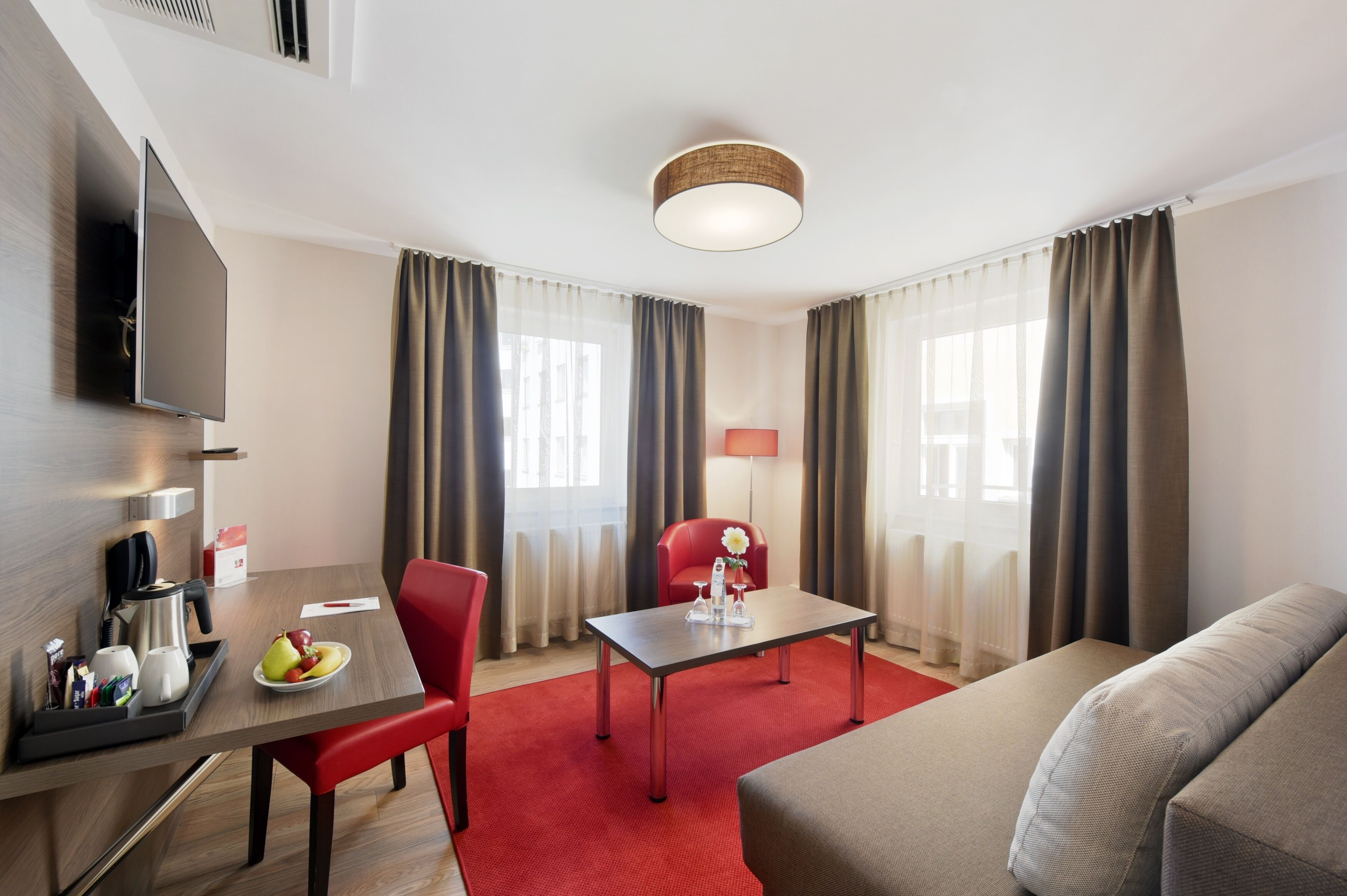 A hotel room with a flat-screen TV, a dining table with a red chair, a sofa, and a lamp.