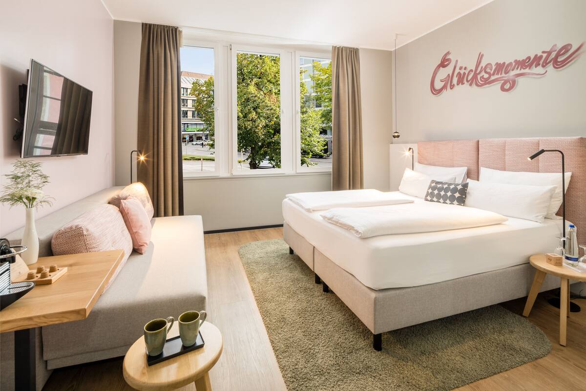 A hotel room with a bed, a sofa, a TV, a window with curtains, a bedside table, and a wall with a sign.