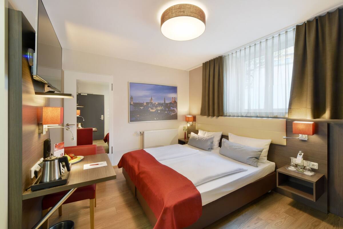 A hotel room with a bed, a desk with a kettle, a chair, a nightstand, and a window with curtains.