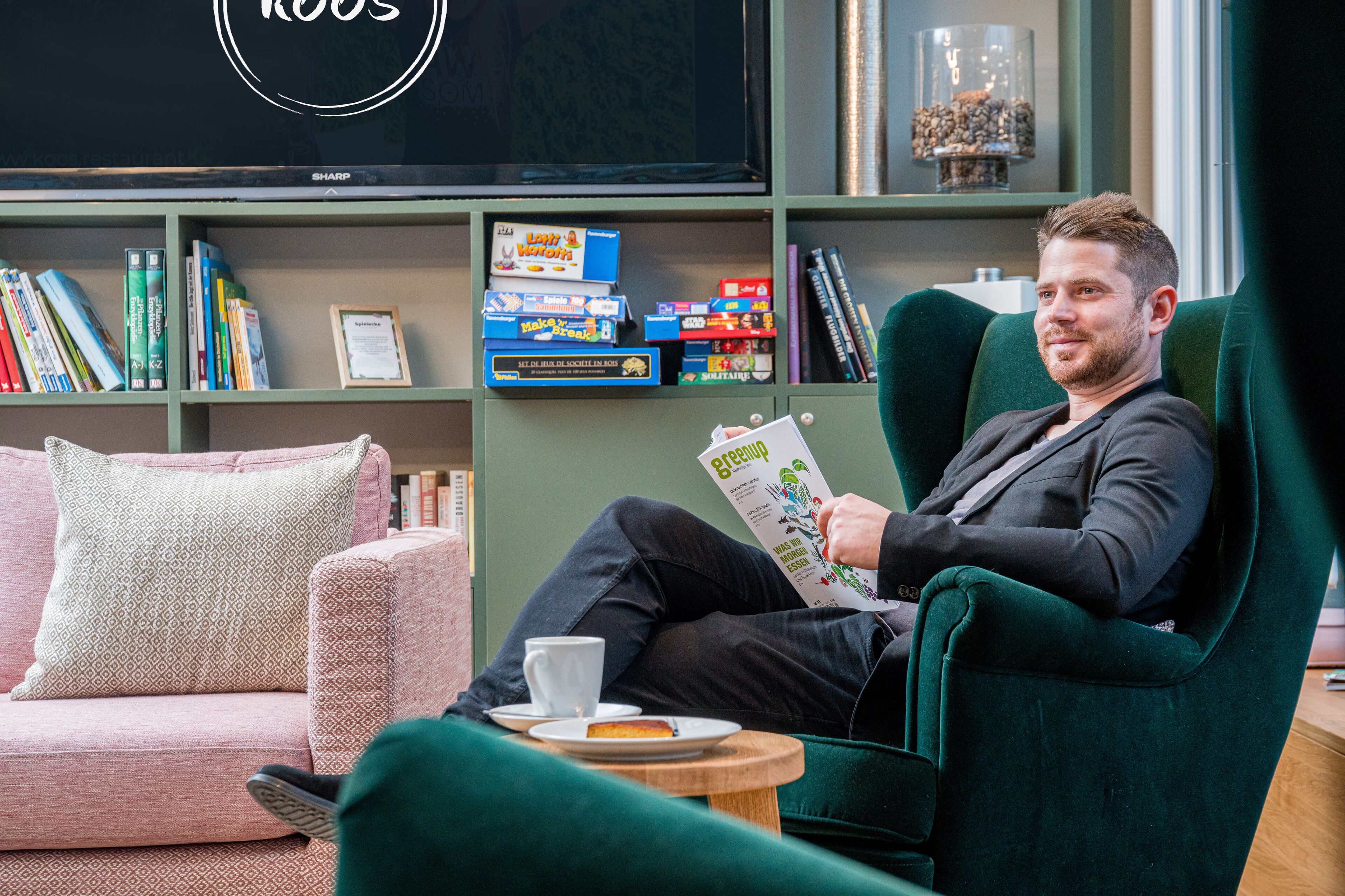 Un homme est assis dans un fauteuil vert, lisant un magazine dans une pièce où se trouvent une bibliothèque, une table basse avec une tasse et une soucoupe, et un canapé rose.