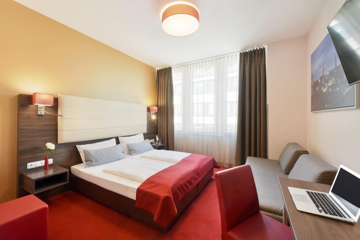 A modern hotel room with a large bed, a desk with a laptop, a red sofa, and a cityscape view.