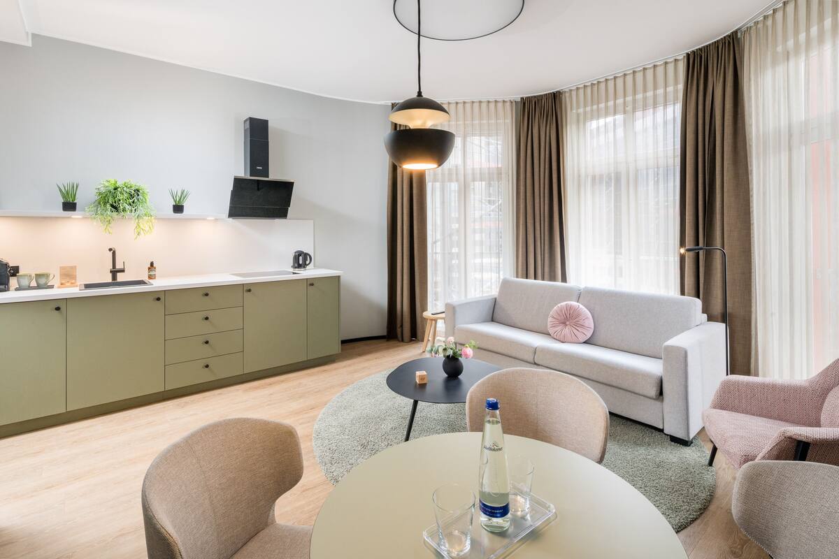 A modern living room with a sofa, a dining table with chairs, a kitchen area with a sink and cabinets, and a large window with curtains.