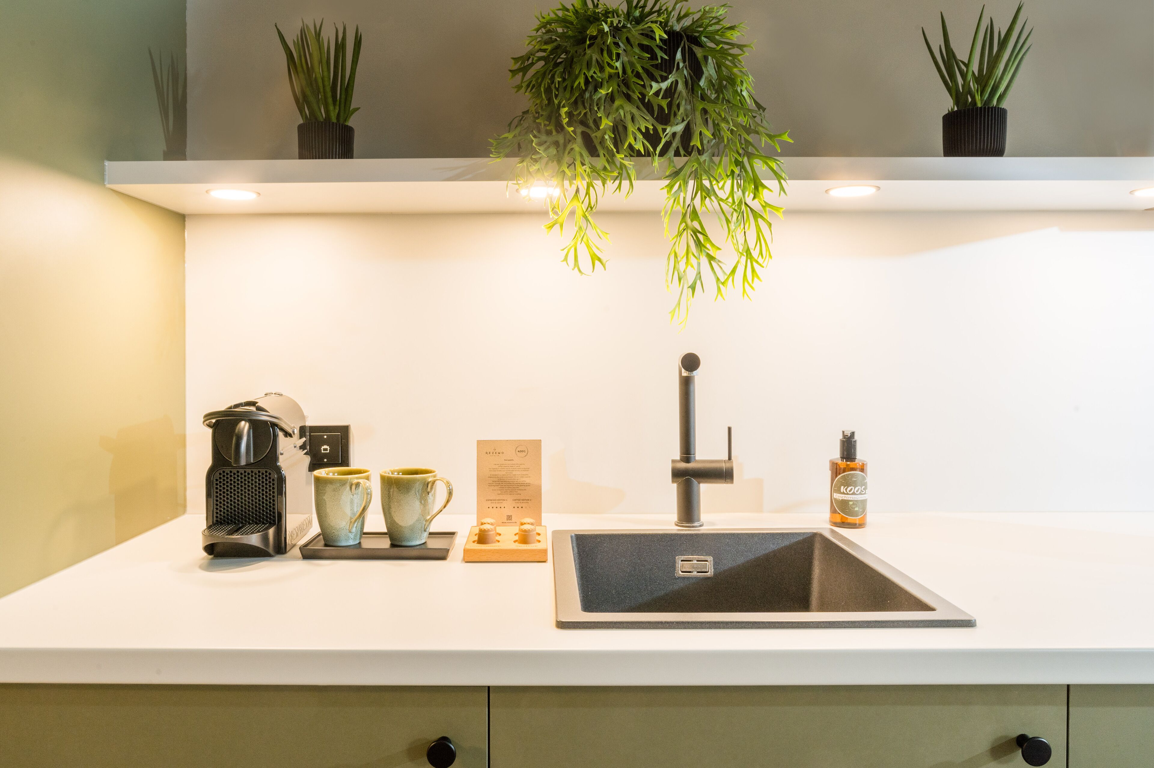 Una cocina moderna con encimera blanca, pileta de acero inoxidable, cafetera, dos tazas, un menú y una botella de jabón para manos.