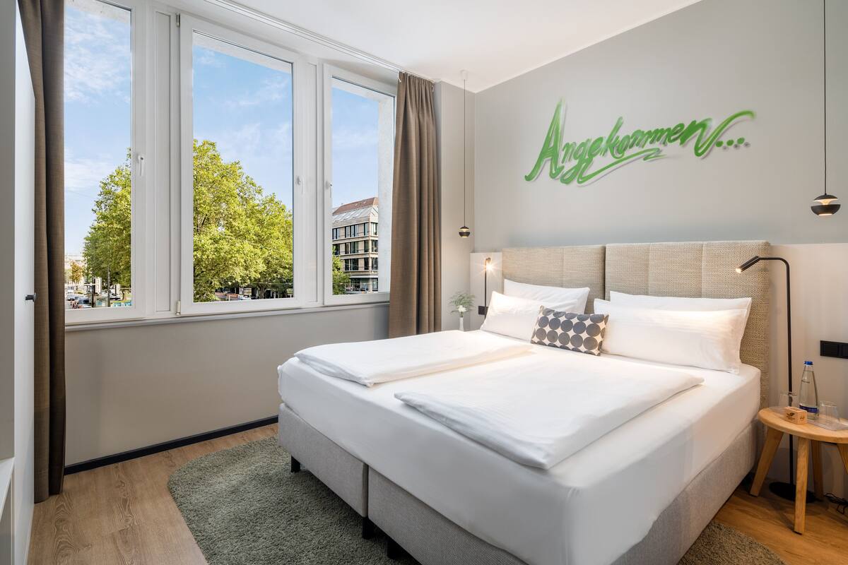 A neatly made bed with white linens, a view of trees and buildings through large windows, and a wall with a green sign.
