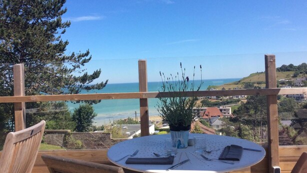 A wooden deck with a round table set for dining, overlooking a coastal view with houses and greenery. - Auberge du vieux puits (Dieppe)