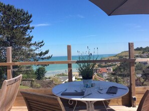 A wooden deck with a round table set for dining, overlooking a coastal view with houses and greenery. - Auberge du vieux puits (Dieppe)