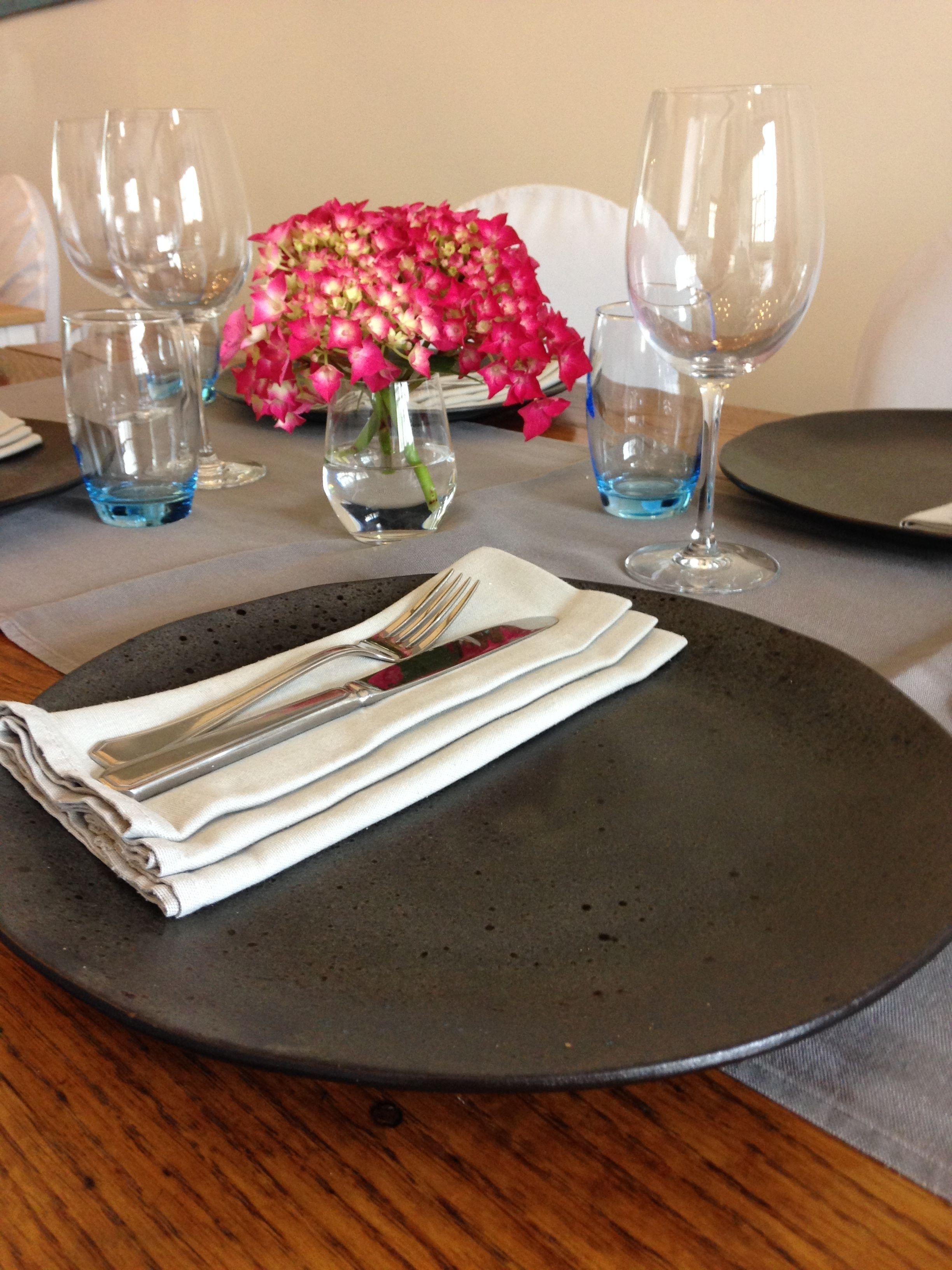 Una mesa dispuesta con un plato oscuro, una servilleta doblada y cubiertos, junto a dos vasos y un jarrón de flores rosadas.