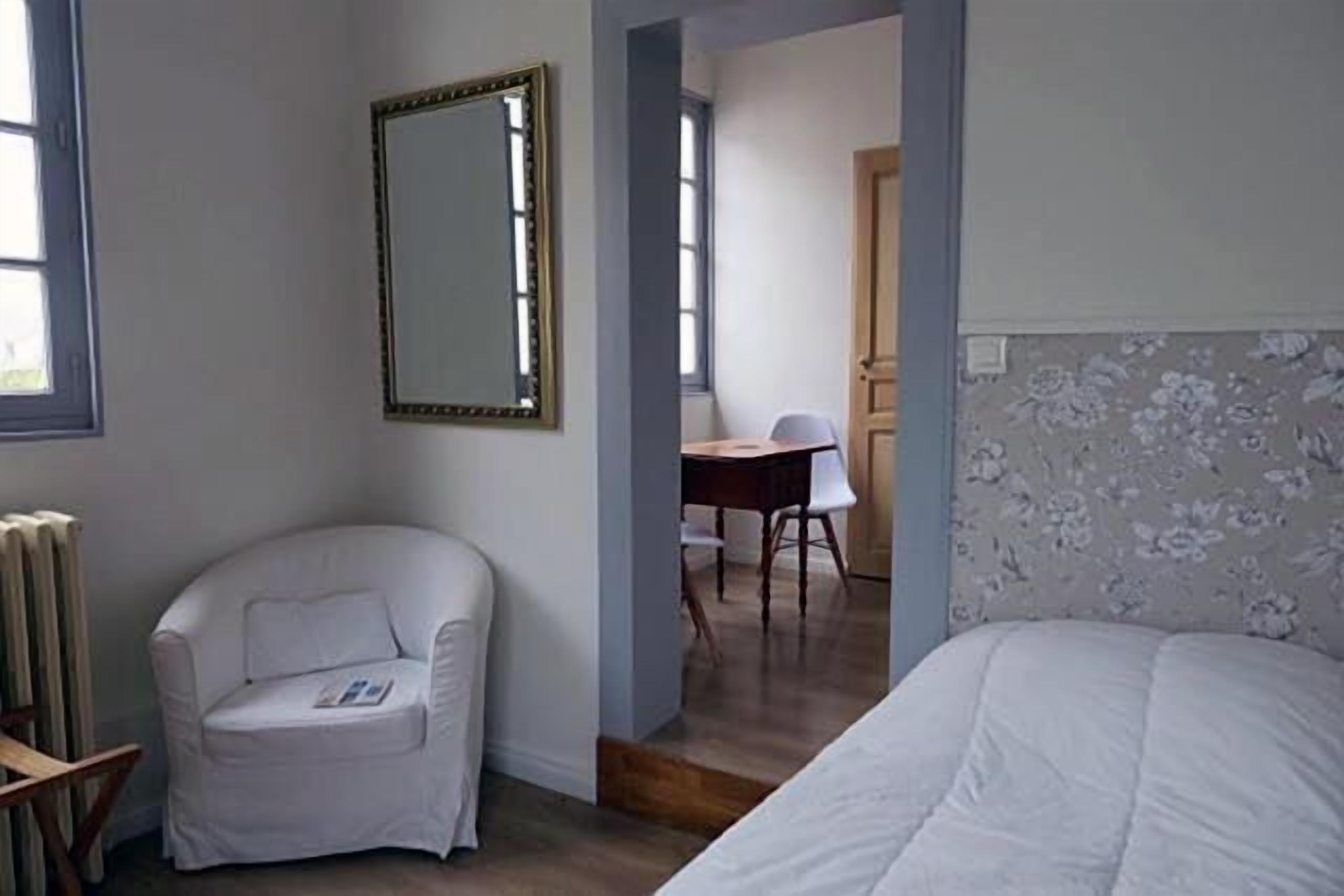 A room with a bed, a white armchair, a desk with a chair, and a large mirror on the wall.