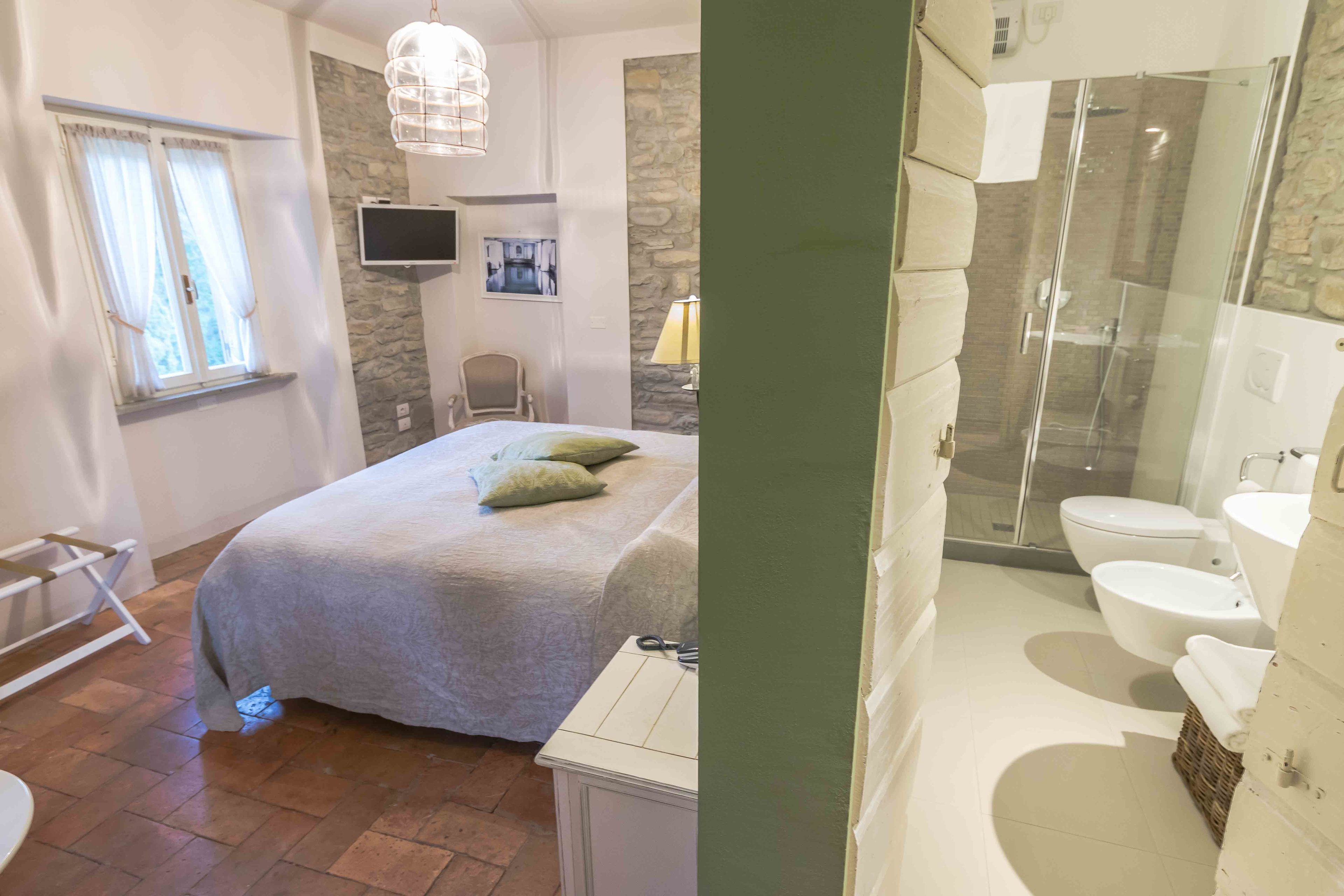 A bedroom with a bed, a chair, a TV, and a window. The bathroom has a shower, a toilet, and a sink.