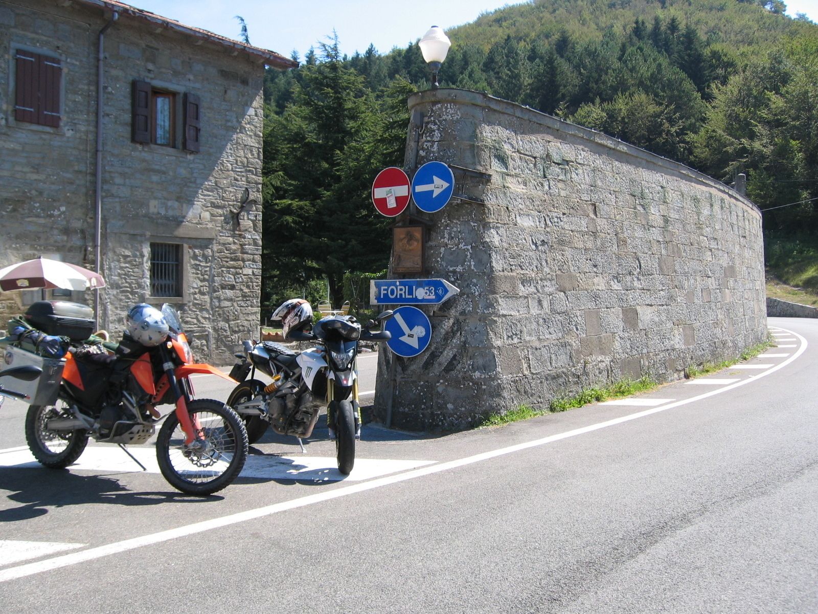 A stone building with a motorcycle parked in front, a 'no entry' sign, and directional signs for 'FORLI' and 'FORLI 53'.