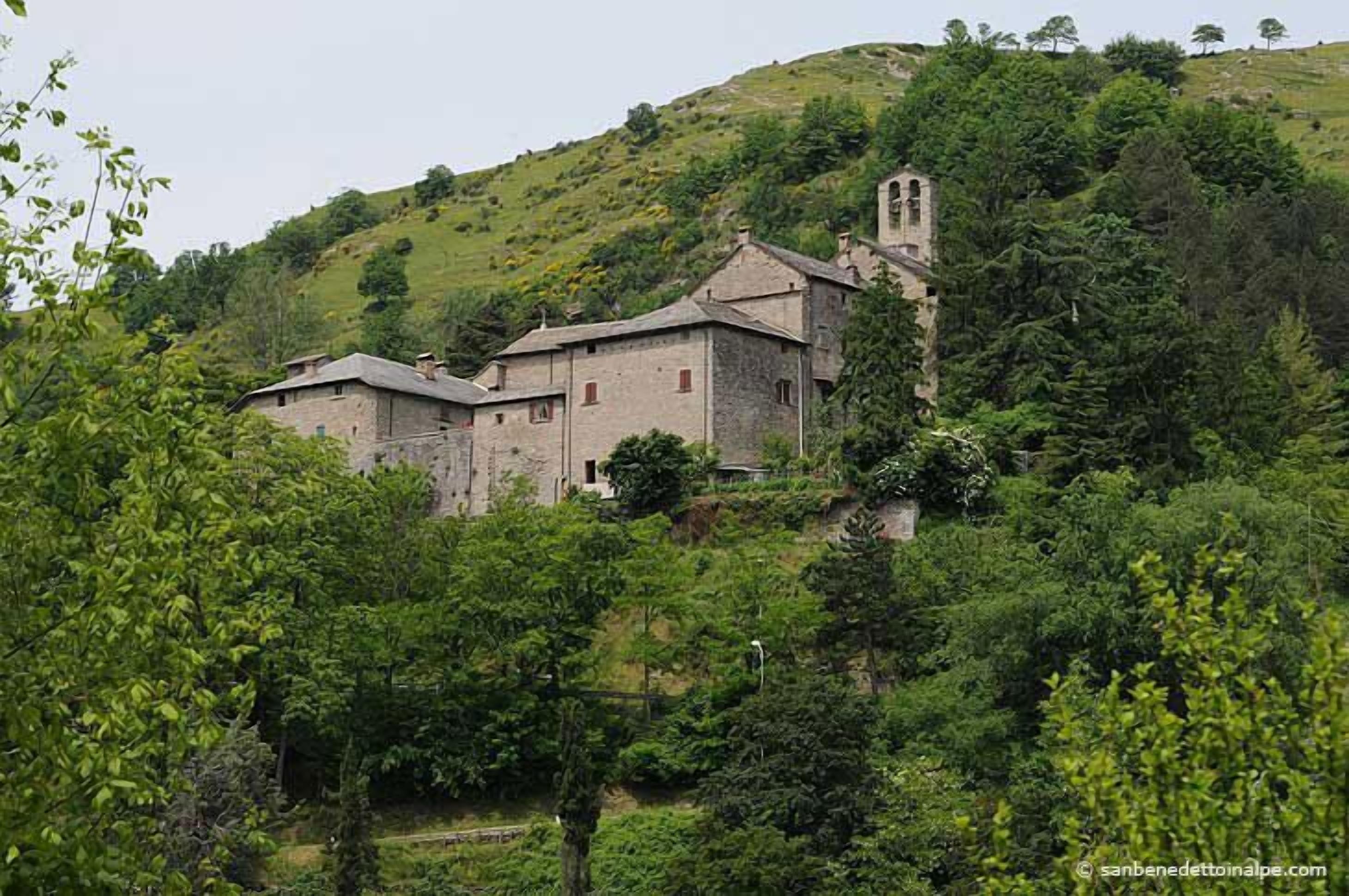 A stone building with a bell tower, surrounded by lush greenery and hills.
